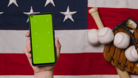 Person-Holding-Green-Screen-Mobile-Phone-Above-Baseball-Still-Life-With-Bat-Ball-And-Catchers-Mitt-On-American-Flag